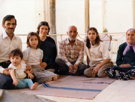 Three children of varying ages, a middle age couple, and an elderly couple sit together in front of large windows which let in significant natural light