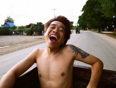 Young shirtless Asian man with dyed light red hair smiling broadly while on the bed of a moving truck on the road