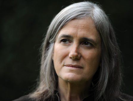 A portrait of a silver-haired woman's face in front of a black backdrop. She looks past the camera