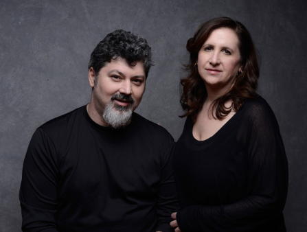 Horizontal portrait of two people from the waist up in front of a grey backdrop. On the left is a man in a long sleeve black shirt with a grey beard and short, dark, curly hair. On the right is a woman in a long sleeve black shirt with long, dark hair.