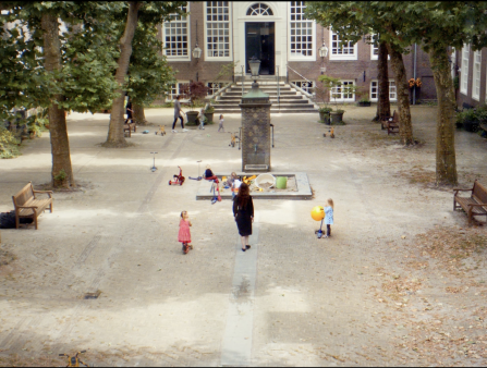 A woman in black walks towards a sandbox in the center of a courtyard. Four little kids and one more woman appear in the courtyard. Kid's scooters and bikes are strewn across the courtyard. There is a row of trees on both sides
