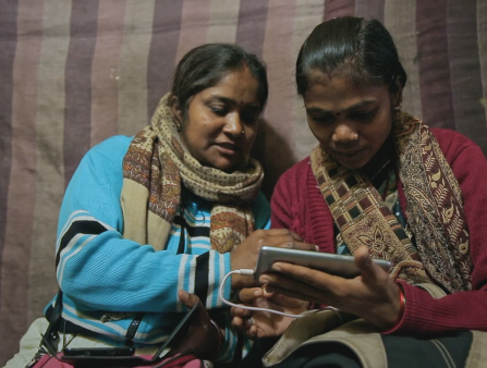 Two woman, one in blue and one in red, both look at a phone held horizontally by the woman on the right (in red). Both women wear brown scarves around their necks and the background is a brown striped fabric.