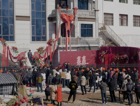 A bride and groom stand on the steps of a large white building decorated in large red bows, banners, and flowers. A crowd of people in winter coats is gathered to observe