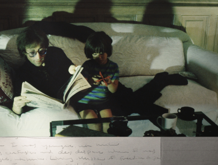 A man in all black reclines on a couch reading while a child in stripes sits next to him