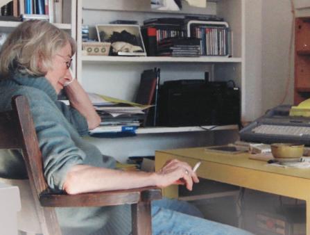 An older woman with grey hair in a turquoise sweater with the sleeves rolled up sits in a wooden chair facing a yellow desk. There is a typewriter and an ashtray on the desk, and she holds a cigarette or joint in her right hand. There is a shelf in the background with books and CDs
