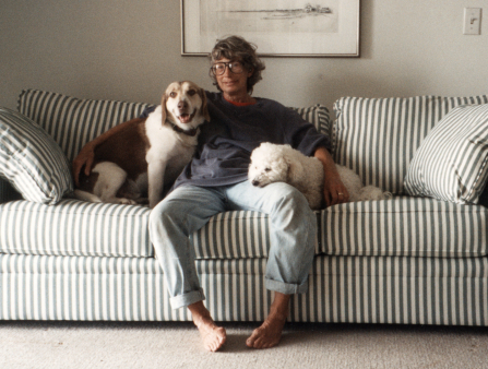 An older woman with short grey hair, glasses, a blue sweater, and cuffed jeans sits barefoot on a striped couch smiling at the camera. There is a framed illustration of a beach house behind her. She holds two dogs, on her right a brown and white medium sized dog smiling at the camera, and on her left a smaller white, curly haired dog resting its head on her lap