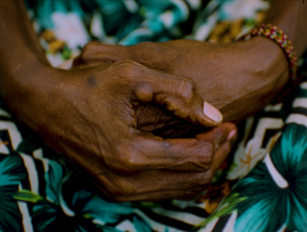 Close up shot of clasped hands resting on green floral fabric. There is a red, yellow, and green bracelet around the wrist of the left hand