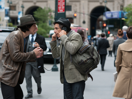 Two men stare intently at each other on a busy sidewalk. On the left, a man in a brown fedora stye hat and brown leather jacket with shoulder length dark hair (Adam Driver). On the right, a man in a grey suit jacket and a grey fedora style hat with a bag over his shoulder holding an audio device to his ear (Ben Stiller) which is connected to a wire held in the hand of the other man