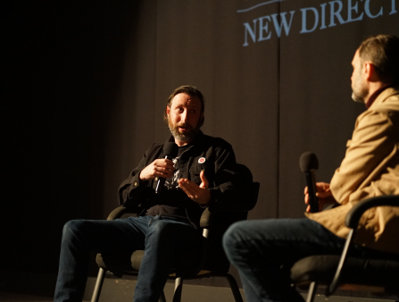 Two men sit in chairs on a stage, each holding a microphone. The man on the left (Megaro) is mid-speech and gestures with his left hand. He wears jeans, a black shirt and a black jacket and boasts a brown beard and mid-length brown hair. The face of the man on the right cannot be seen. He wears a beige jacket and jeans