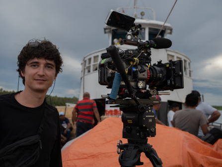 A young man in a black shirt with short, dark, curly hair smiles beside a large camera rigged with a microphone, monitor, and other equipment on top of a tripod. The man and camera are on a boat and passengers, the bridge of the ship, and a glimpse of the water can be seen behind them