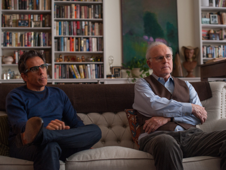 Two men sit side by side on a white couch in front of a wall of bookshelves. On the left is a man in a blue sweater and glasses with his legs crossed (Ben Stiller), and on the right is an older man, also wearing glasses along with a brown vest over a light blue collared button down shirt