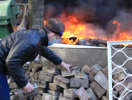 A man in a leather jacket and mask places a brick on a pile in front of burning flames and a green dumpster