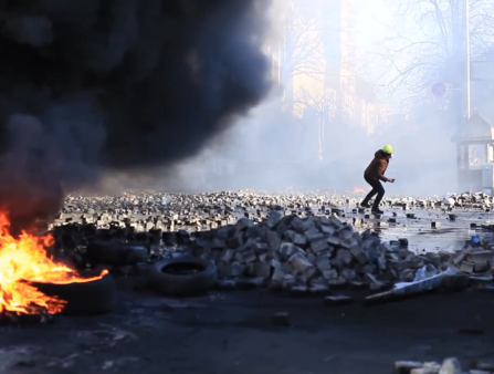 A man stands amidst piles of bricks preparing to throw something. In the foreground, a fire burns and two people observe