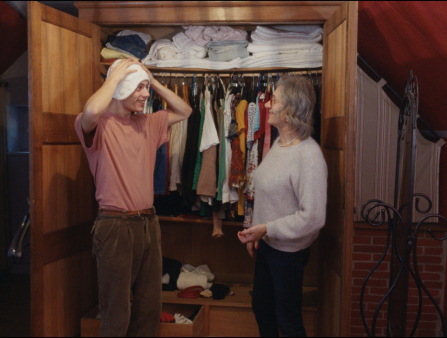 Two people stand in front of a full wardrobe. On the left is a young man wearing a red T-shirt tucked into brown pants, holding a white towel on his head. On the right is a woman with grey hair, glasses, a grey sweater and black slacks. The two smile at each other