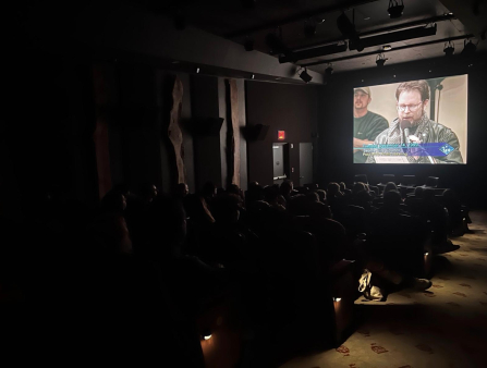 A theatre full of people look at a screen which displays a man in a leather jacket and glasses speaking into a microphone. Text on the screen reads "Tuesday December 14, 1999, Seattle City Council, Post-WT Public Hearing"