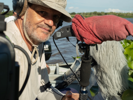 A bearded man sits on a boat and smiles, wearing headphones over a bucket hat and holding a microphone on a boom pole