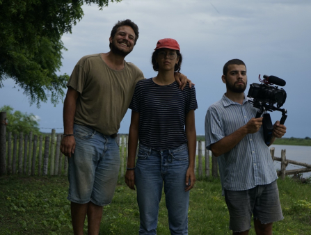 Three people stand side by side. On the left, a man in a green T-shirt and jean shorts smiles widely while holding the shoulder of the woman in the center, who wears glasses, a red hat, jeans and a striped shirt. On the right a man in a short-sleeved light blue button-down collared shirt holds a camera with a microphone mounted on top