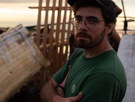 A man in a green T-shirt, black cap and round glasses with brown facial hair crosses his arms and looks to his left into the camera