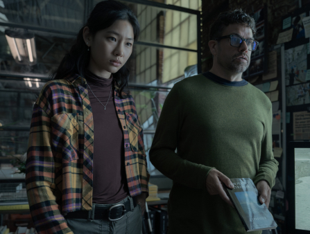 A woman on the left in a flannel shirt over a red sweater and a man on the right holding a book and wearing glasses and a green sweater stare at something off camera in shock. The background is a large warehouse with windows letting in yellow light