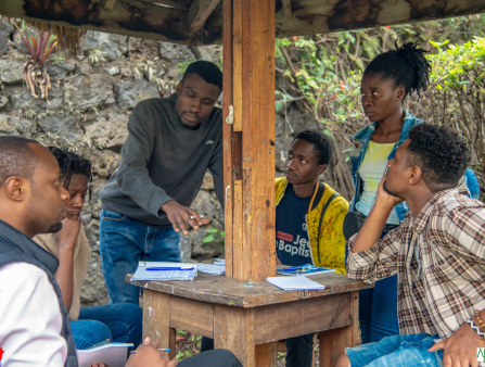 Six people gather around a small wooden table with a wooden column rising from its center supporting a wooden roof. Four sit and two stand. One of the two standing is a man in a grey turtleneck and blue jeans, gesturing emphatically. The rest watch and listen. Notebooks and pens sit on the table. A rock wall and vegetation fill the background