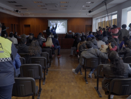 Shot from the back, a room full of people in chairs stare at a screen on the far wall. Some stand. In the back stands a woman in a yellow and blue jacket which reads "POLICIA." On the right of the room someone holds a microphone on boom pole. The screen displays an image of a man walking. Someone stands in front of the screen and points at it