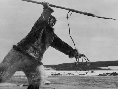 Black and white photo of a man in a fur coat holding a spear above his head amidst a sparse, icy background