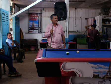 A man in a pink collared button down shirt holds a pool cue and stands at a pool table with a blue top and an image of the eiffel tower on the side. He is in a white building with blue doors. Half a dozen people sit around the room and two sit outside, seen through open doors. Another pool table and a large speaker are behind the man