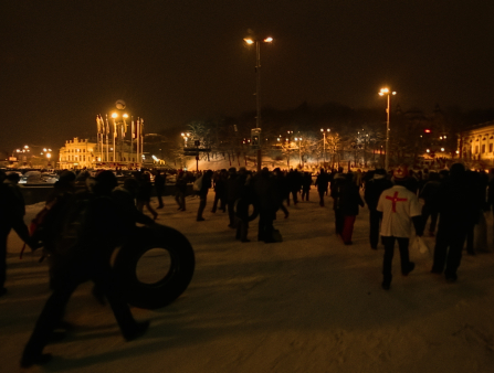A large crowd marches through an open space at night. Tall streetlights and flags of various countries can be seen