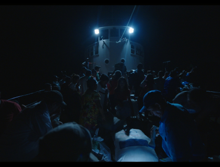 Night time shot of a crowd of people gathered on a boat. Two blue lights on top of the large bridge are the only source of light