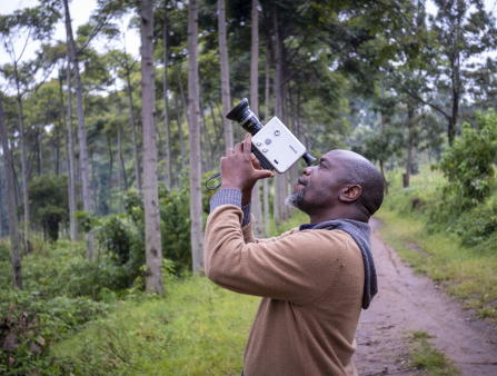 A Black man in a beige sweater over a grey sweater stands on a path beside a number of trees, looking through the viewfinder of a small, white camera he points to the sky