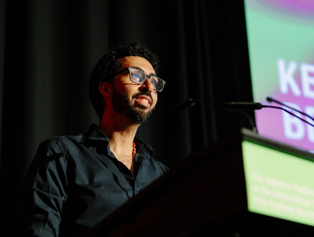 A man in a black shirt with black glasses and short dark hair and beard delivers a speech at a podium. Behind him is a black curtain and the lefthand side of a pink and green digital display showing text which cannot be read