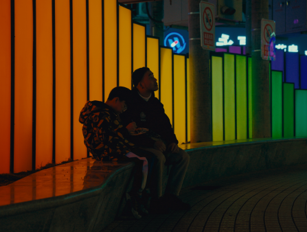 An Asian man and a young boy sit at night in front of a rainbow-lit public display