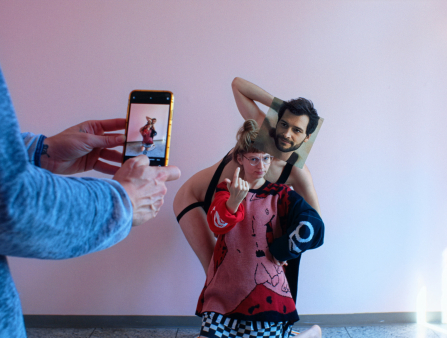 A woman wearing a red dress with a drawing of a naked man printed on it sits in front of someone almost naked with anothther face photoshopped over theirs. On the left, two hands holding a cell phone can be seen documenting the pair