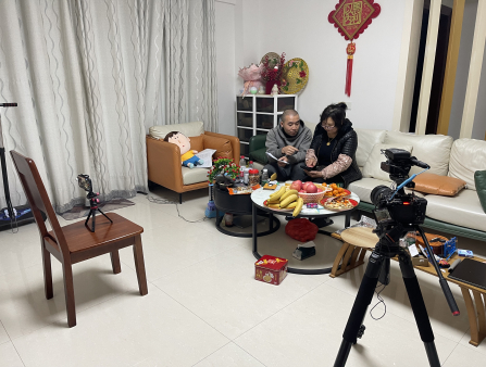 A camera on a tripod stands in the foreground as an Asian man sits down next to an Asian woman in the middle of their living room