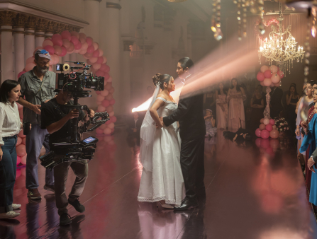 In the center of the frame, a couple in wedding dress and tuxedo dance. On the left, a woman and a man stand behind another man with a steadicam filming the two. On the right, a crowd of wedding attendees stand in colorful, nice clothes