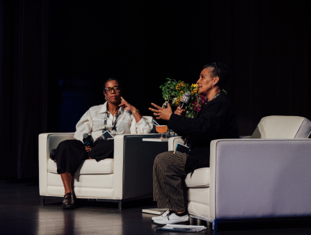 Two black woman sitting in oversized couch chairs on stage holding mics addressing one another