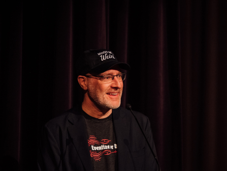 A middle-aged man with salt and pepper stubble wears a cap that reads "Movies Love Weird" and a t-shirt under a blazer that reads "Eventize or Die"
