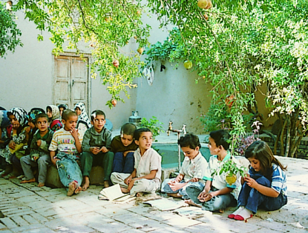 A large group of children sit under a tree in a sunlit stone-paved courtyard