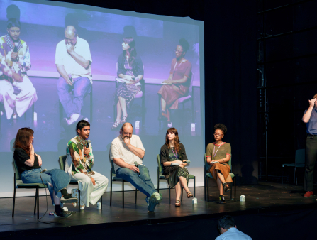 Three women and two men sit in simple chairs on a stage and hold microphones. The five people are also displayed live on a large screen behind them all. On the right, a man in black slacks and a blue button-down shirt stands and signs