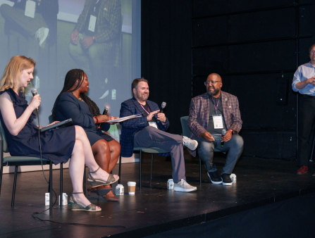 Two women in dark blue dresses and two men in plaid suit jackets sit in a row of chairs on a stage, with a large screen behind them displaying their image. A man in black slacks and a blue button-down shirt stands behind them, signing