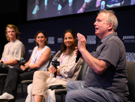 A row of four people sit in chairs and hold microphones on a stage. The three in the background, one young man with long hair and two young women, are out of focus, and the person in the foreground is a middle-aged man gesturing with his left hand