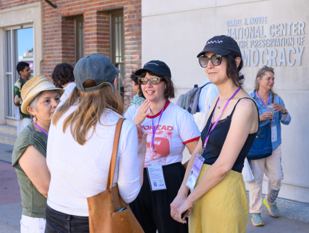 For women wearing lanyards stand in a circle mid-discussion. Several other people walk behind them. The brick building they stand in front of displays text which reads "DANIEL K. INOUYE NATIONAL CENTER FOR THE PRESERVATION OF DEMOCRACY"