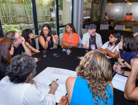 Eleven people sit around a round table covered in a black tablecloth, one of whom writes with marker on a large white poster. In the background are more round tables covered in black tablecloths and large windows which show a space decorated with stones and bamboo