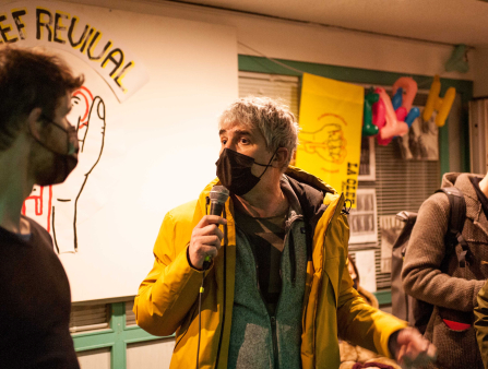 A man with short grey hair wearing a large yellow coat and a black medical mask faces a young man, also masked, and speaks into a microphone. They are in a room the walls of which are lined with posters and art