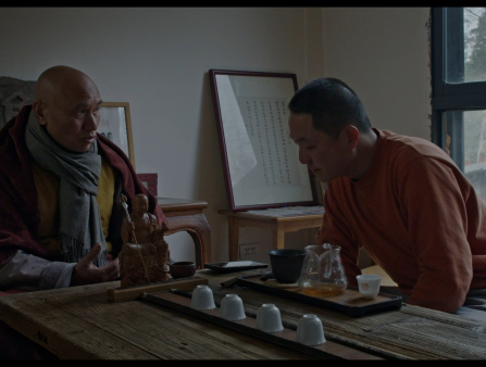 A bald man in robes and a scarf sits across from a man with a buzzcut in an orange shirt at a low table. On the table are a wooden sculpture of a monk, a pitcher, and four teacups. In the background is a large window and two tables displaying art, including a framed piece of calligraphy