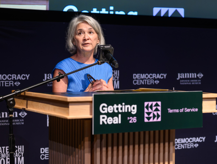 A woman in a blue dress stands at a podium which reads "Getting Real '26" and speaks into a microphone