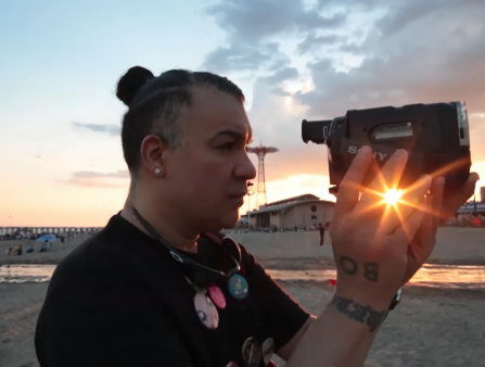 A person with a topknot looks into the viewfinder of a Sony camcorder on the beach at sunset. They wear a black T-shirt dotted with pins and a diamond stud earring. The sun shines through their fingers