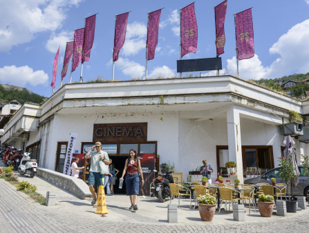 Two people walk out of a white building which displays a sign that reads 'CINEMA.' 9 pink banners fly from the roof of the building and yellow tables and chairs sit in front