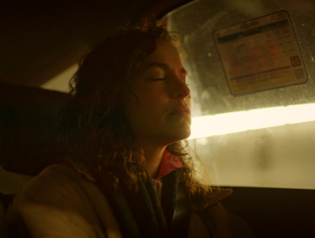 Close-up of a woman sitting in the backseat of a car with her eyes closed. The car is passing through a tunnel. The woman has curly hair and wears several layers.