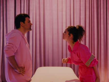 A man and a woman, both dressed fully in pink, stand across from each other. There is a massage table in between the two, and the background is a pink curtain. The woman, on the right, yells at the man, who stands on the left with his hands on his hips